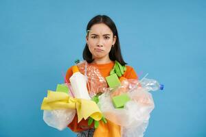 Upset girl has to recycle, holds plastic bottles and litter, looking bothered and angry, blue background photo