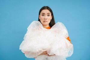 Annoyed woman holding loads of plastic in hands, looking bothered with sorting trash, eco-activist fighting for nature preservation, blue background photo