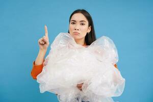 Asian woman with plastic bags, recycling waste in hands, points up, showing recycle sorting center, sustainable life tips, blue background photo