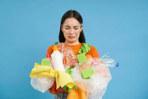 ceñudo asiático mujer con el plastico basura en manos, quejumbroso a no reciclaje desperdiciar, en pie con camada terminado azul antecedentes foto