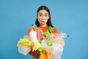 Young confused asian girl with different types of plastic, learns how to recycle, saving enviroment, sorting garbage, blue background photo