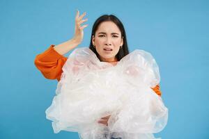 Annoyed asian woman volunteer, holding plastic waste, shouting with disappointed face, hates polution, stands over blue background photo