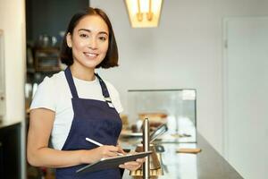 Small cafe business. Smiling asian girl barista in apron, using tablet as POS terminal, processing coffee order behind counter photo