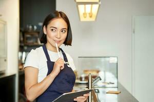 Smiling cute barista girl, korean woman working in cafe, using tablet to process order, making coffee at counter photo