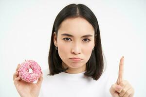 Close up of serious asian woman, shows taboo, stop gesture and pink glazed doughnut, forbids eating sweets, white background photo