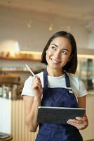 Vertical shot of smiling girl waitress, barista in coffee shop, wears blue apron uniform, takes order with tablet, stands in cafe photo