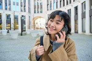 celular tecnología. joven coreano mujer negociaciones en móvil teléfono, hace un teléfono llamada en su camino hogar, camina abajo calle, ciudad centro, tiene teléfono conversacion foto