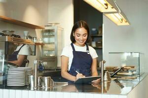 Smiling cute barista girl, korean woman working in cafe, using tablet to process order, making coffee at counter photo