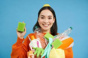 Enthusiastic asian girl showing cleaning sponge, holding empty plastic bottles, trash for recycling, sorting her household garbage, blue background photo