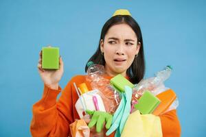 retrato de asiático mujer mirando disgustado a sucio esponja, participación reciclable vacío botellas, clasificación basura, azul antecedentes foto