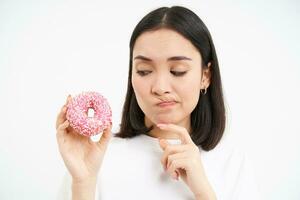 cerca arriba de niña piensa de frenado su dieta, mirando a tentador vidriado rosquilla, blanco antecedentes foto