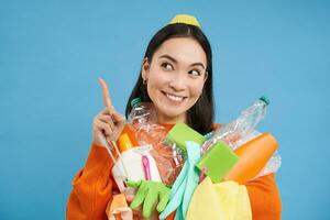 Enthusiastic asian woman, holding plastic empty bottles and garbage for sorting and recycling, pointing finger up at recycle center, blue background photo