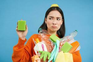 Portrait of asian woman looking disgusted at dirty sponge, holding recycable empty bottles, sorting garbage, blue background photo