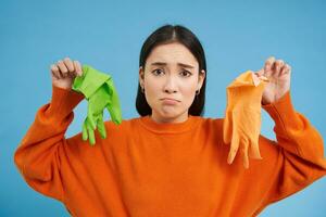 Portrait of sad korean woman, showing two latex gloves with upset face, dislikes cleaning, standing over blue background photo