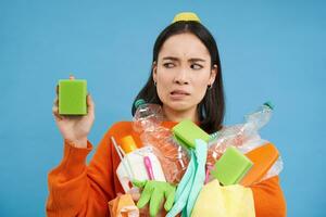 Portrait of asian woman looking disgusted at dirty sponge, holding recycable empty bottles, sorting garbage, blue background photo
