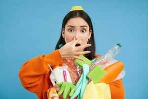 Portrait of woman shuts her nose from digust, holding stinky garbage, dirty plastic items and empty bottles with bad smell, standing over blue background photo