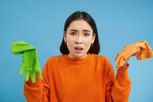 confuso asiático mujer, demostración dos látex guantes, se queja a limpieza hogar, en pie terminado azul antecedentes foto