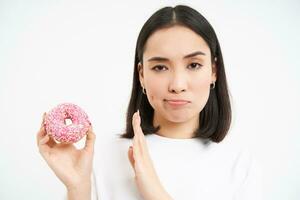 No sweets. Serious young woman on diet, showing stop gesture and glazed doughnut, disapprove junk food, white background photo