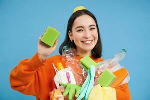 Smiling enthusiastic female eco activist, holding cleaning sponge, empty plastic bottles and garbage, sorting waste for recycling, blue background photo