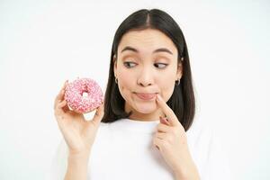 Smiling young korean woman, thinking and gazing at pink glazed doughnut, white background photo