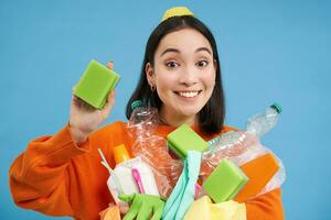 Smiling enthusiastic female eco activist, holding cleaning sponge, empty plastic bottles and garbage, sorting waste for recycling, blue background photo