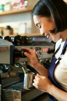 contento asiático mujer, barista utilizando café máquina a hacer orden, humeante Leche para capuchino y latté, riendo y sonriente mientras trabajando en café foto