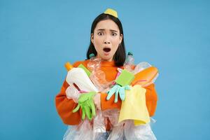 Sad asian woman holding garbage, empty plastic bottles and waste, tired of recycling, sorting rubbish, isolated on blue background photo