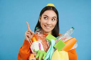 Enthusiastic asian woman, holding plastic empty bottles and garbage for sorting and recycling, pointing finger up at recycle center, blue background photo