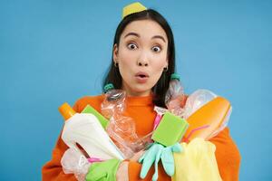 Close up of girl with surprised face, holds empty plastic bottles and green waste, collects garbage for recycling, blue background photo