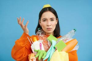 Portrait of woman puzzled by recycling rules, holding empty plastic bottles and garbage, looks complicated, blue background photo