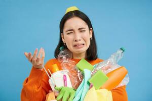 Crying korean woman complaining, holding garbage, plastic bottles, litter, sorting garbage, stressed by recycling, blue background photo