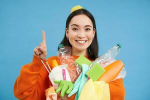 Portrait of smiling korean woman, pointing up, recycling waste, holding plastic garbage and looking happy, blue background photo