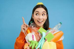 Enthusiastic asian woman, holding plastic empty bottles and garbage for sorting and recycling, pointing finger up at recycle center, blue background photo