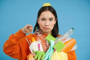 Serious korean woman pointing finger at camera, blame people for garbage polution, holding empty plastic bottles and waste, telling to recycle, blue background photo