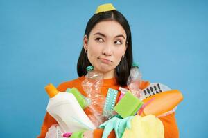 Portrait of asian woman with empty plastic bottles, looks aside with thinking face, recycle garbage at home, blue background photo