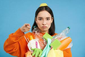 Serious korean woman pointing finger at camera, blame people for garbage polution, holding empty plastic bottles and waste, telling to recycle, blue background photo