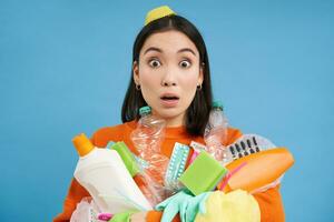 Portrait of girl with shocked face, holds empty plastic bottles, household garbage, stares startled at camera, recycles, blue background photo