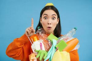 Close up portrait of asian woman pointing up, showing banner, holding recycling items, empty plastic bottles, sorting garbage, isolated on blue background photo