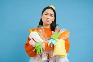 Sad asian woman holding garbage, empty plastic bottles and waste, tired of recycling, sorting rubbish, isolated on blue background photo