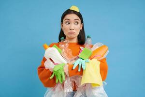 Confused young asian woman, holding plastic bottles, household garbage, sorting waste for recycling, blue background photo