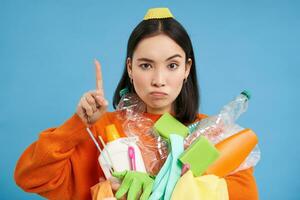 Close up portrait of asian woman pointing up, showing banner, holding recycling items, empty plastic bottles, sorting garbage, isolated on blue background photo