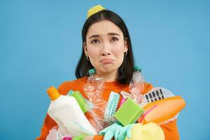 Portrait of crying sad girl with plastic trash, collects recycling waste, looks miserable, blue background photo