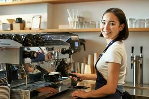 sonriente asiático barista niña hace capuchino con café máquina, soportes detrás mostrador en café foto