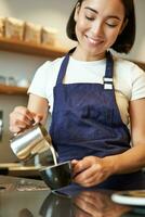 Vertical shot of brunette asian girl barista, wearing apron, making coffee, prepare order in cafe, pouring milk in cup for cappuccino latte art photo