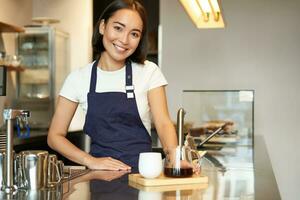 sonriente hermosa barista niña en delantal, haciendo lote elaborar cerveza, filtrar café, en pie en café detrás mostrador foto