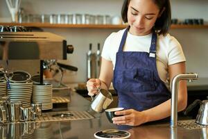 Happy smiling cafe owner, girl barista in apron, making cappuccino, latte art with steamed milk, standing behind counter photo