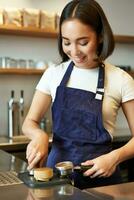 Vertical shot of smiling asian girl putting coffee in tamper to brew capuccino, prepare order in coffee machine, standing behind counter in blue apron photo