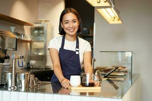 sonriente hermosa barista niña en delantal, haciendo lote elaborar cerveza, filtrar café, en pie en café detrás mostrador foto