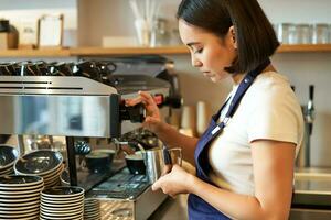 Smiling young asian barista girl, using coffee machine steamer, steaming milk for cappuccino order, standing in apron, working in cafe photo