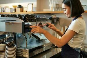 Smiling cute korean girl, barista working with coffee machine, prepare cappuccino, using steamr at cafe counter photo